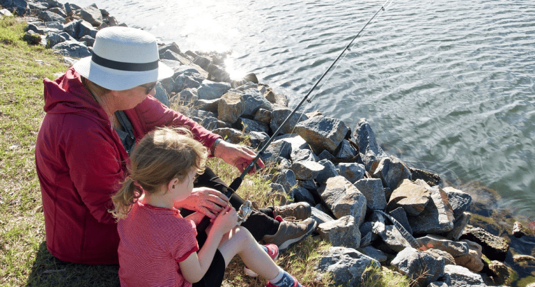 Relaxing outdoor scene of a grandmother and young girl fishing by the lake, enjoying nature and quality time together. Peaceful moment captured with scenic water and rocks in the background.