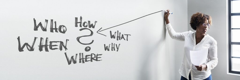 A woman of color draws with a marker on a white board while facilitating a class. Who, What, Why, Where, and How are written on the board in a diagram