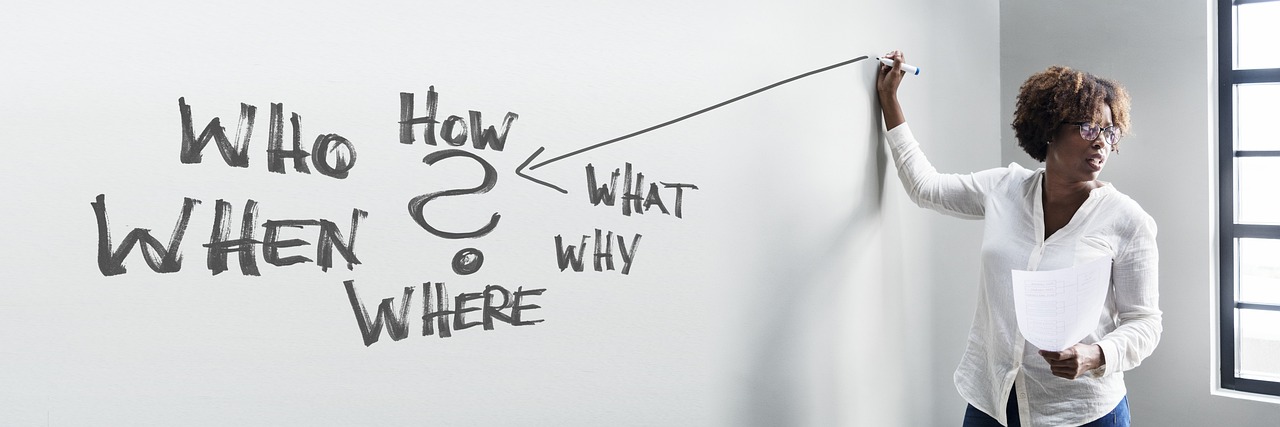 A woman of color draws with a marker on a white board while facilitating a class. Who, What, Why, Where, and How are written on the board in a diagram