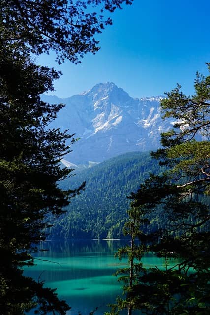 View of a blue, distant mountain peak with spots of snow as seen through the gap of two evergreen trees.