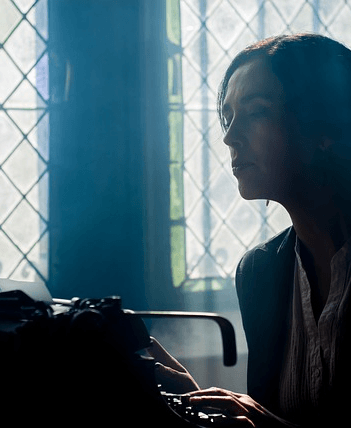 Partial image of a female writer that is in front of a typewriter. A lit window is in the background, making her appear dark as though in a shadow.