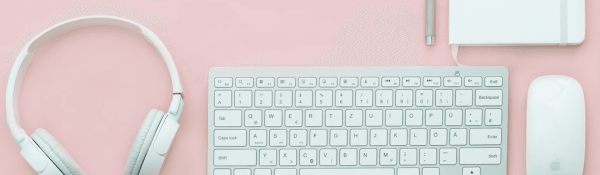 A clean, pink desk top with headphones, a keyboard, mouse and a blank pad of paper.