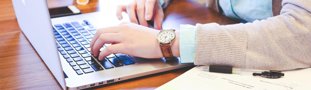 Close up photo of a woman's arm and hands typing on a laptop. She is wearing a brown watch and a pink sweater