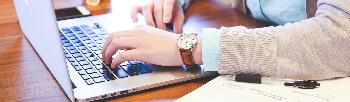 Close up photo of a woman's arm and hands typing on a laptop. She is wearing a brown watch and a pink sweater
