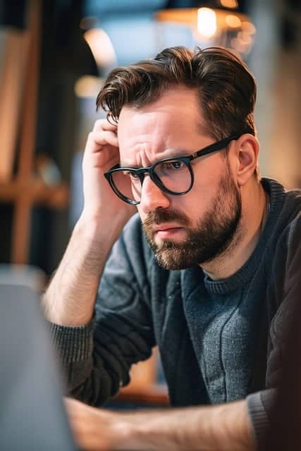 Thoughtful man with glasses working on laptop in a modern office environment, representing focus and problem-solving skills.