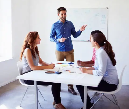 Two women sit at a table while a man stands next to them. A white board is ready for jotting down ideas.