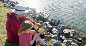 A Grandmother wearing a sunhat helps a youngster learn how to fish at the the water's edge.