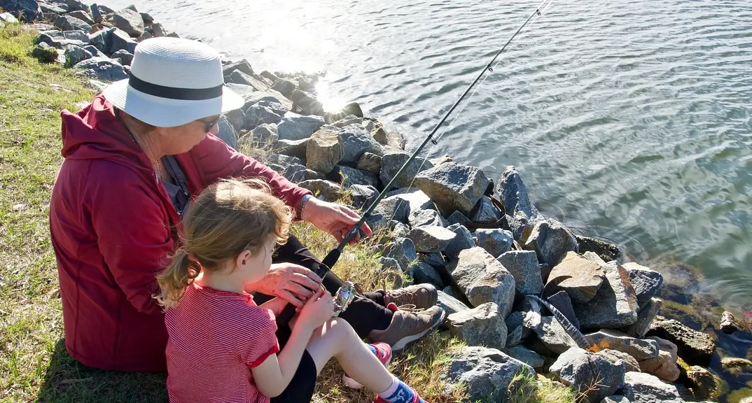 A Grandmother wearing a sunhat helps a youngster learn how to fish at the the water's edge.