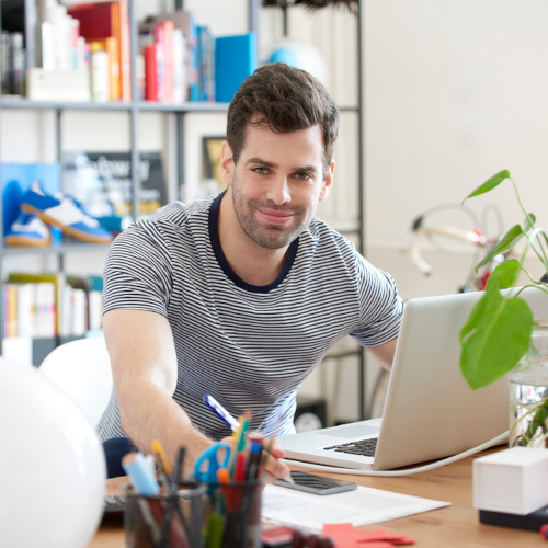 A man sits at the desk in his home office surrounded by plants, writing utencils and a bookcase with colorful items on the shelves.