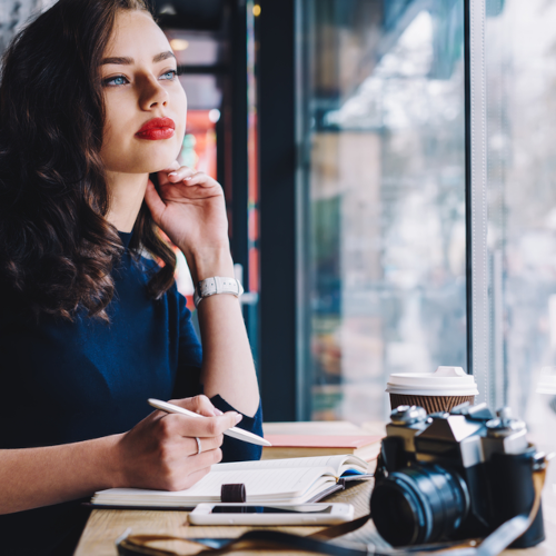 A woman sits thoughtfully, a cup of coffee next to her camera. She is being thoughtful, pen in hand, paper ready