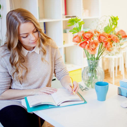 A woman with long blonde hair writes in a planner surrounded by meticulous order and cleanliness