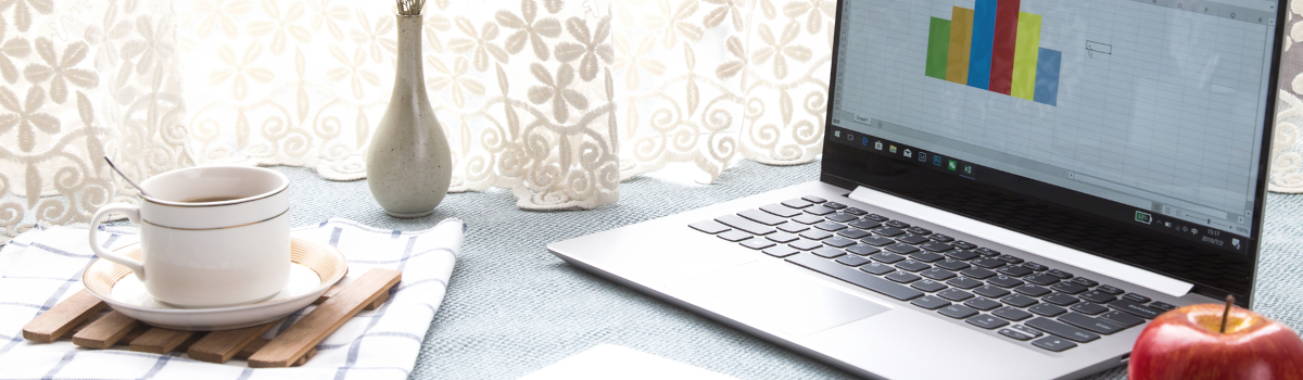 Partial photograph of a laptop that is open. A sales graph is on the screen. Various desk items such as a pad of paper, pen, apple, flower vase are in the image.