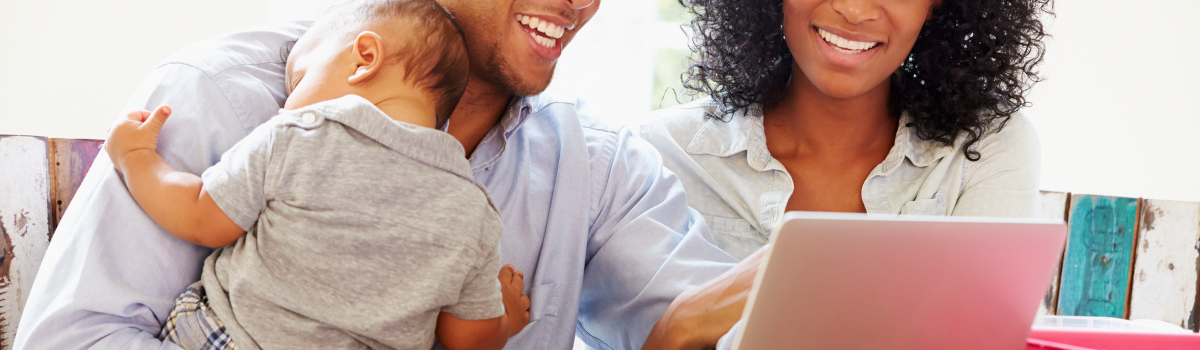 A black father holds his sleeping infant while seated next to his wife. They are looking at their laptop and smiling